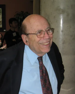Michael O. Rabin 2004: ein älterer Mann mit Halbglatze und Brille in hellblauem Hemd, gemusterter Krawatte und dunklem Sakko, lachend.
