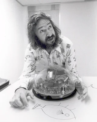 Schwarzweißfoto: Seymour Papert mit langen Locken und Vollbart an einem Tisch, vor ihm ein runder Roboter unter einer durchsichtigen Kuppel und auf dem Papier eine Zeichnung aus geraden Linien.