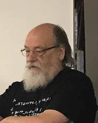 Ken Thompson 2019: ein Mann mit Brille, weißem Bart und Halbglatze in einem schwarzen T-Shirt.