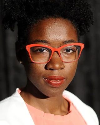 Porträt von Joy Buolamwini vor dunklem Hintergrund, mit lockigem Haar und einer roten Brille, in weißer Jacke über einem lachsfarbenen Oberteil.