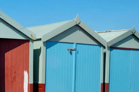 Ein Foto von Strandhäusern: links eine rote Tür mit einem weißen Farbfleck, daneben zwei hellblaue Türen unter blassgrünen Giebeln, darüber blauer Himmel.
