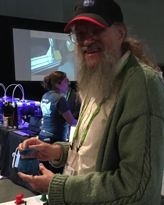Jim Blinn 2015 auf der SIGGRAPH: ein Mann mit langem grauem Bart, Brille und NASA-Mütze in grüner Strickjacke, lachend, in den Händen eine blaue Teekanne aus dem 3D-Drucker.