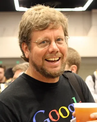 Guido van Rossum 2006: ein lachender Mann mit Brille und Bart in einem Google-T-Shirt, mit einem Becher in der Hand.