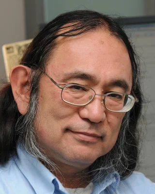 Jim Kajiya 2009 in seinem Büro: ein Mann mit Brille, langem dunklem Haar, das an den Seiten grau ist, und hellblauem Hemd, mit leichtem Lächeln; hinter ihm Bildschirme.