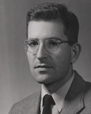 Schwarzweißporträt des jungen Noam Chomsky: ein Mann mit kurzem Haar und runder Brille, in Jackett und dunkler Krawatte, leicht zur Seite blickend.