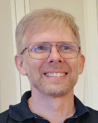 John Carmack 2025: ein Mann mit kurzem blondem Haar, Brille und Dreitagebart in einem dunklen Polohemd, breit lächelnd.